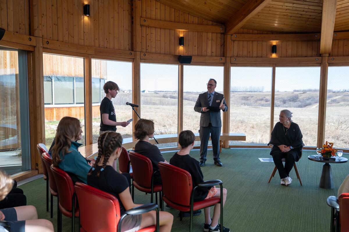 A young person standing, speaking into a microphone. Governor General Simon and others are seated.