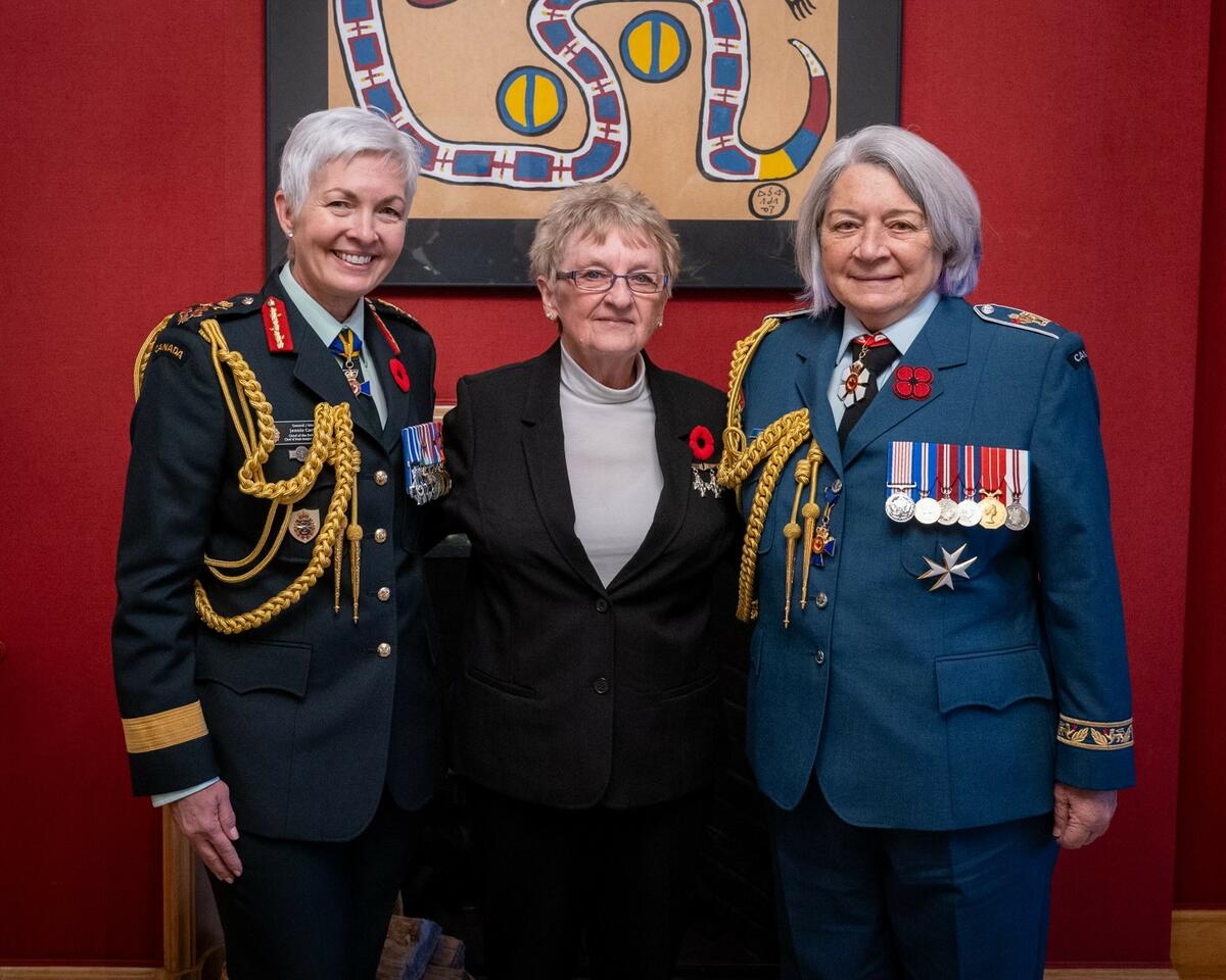 Une femme portant des lunettes est entourée de deux dames en uniforme. À sa gauche, on aperçoit le général Jennie Carignan, chef d'état-major de la Défense du Canada, et à sa droite, la gouverneure générale Mary Simon.