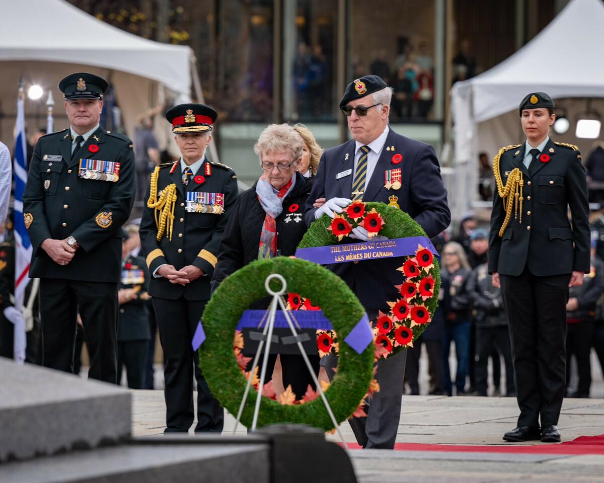 Un militaire aux cheveux gris tient une couronne de fleurs avec un ruban portant l'inscription « The Mothers of Canada ». Une dame à lunettes, portant un foulard bleu clair et rouge autour du cou et inclinant la tête, se trouve à côté de lui.