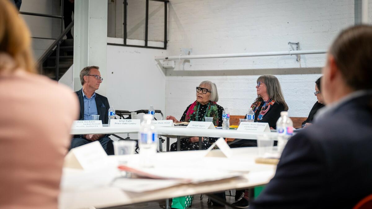 One grey haired lady, with glasses, is speaking, seating at a table corner. A man on her right and a lady at her left as seated on both sides. Their names are on tent cards in front of them. People seen from the back are blurred on either side .