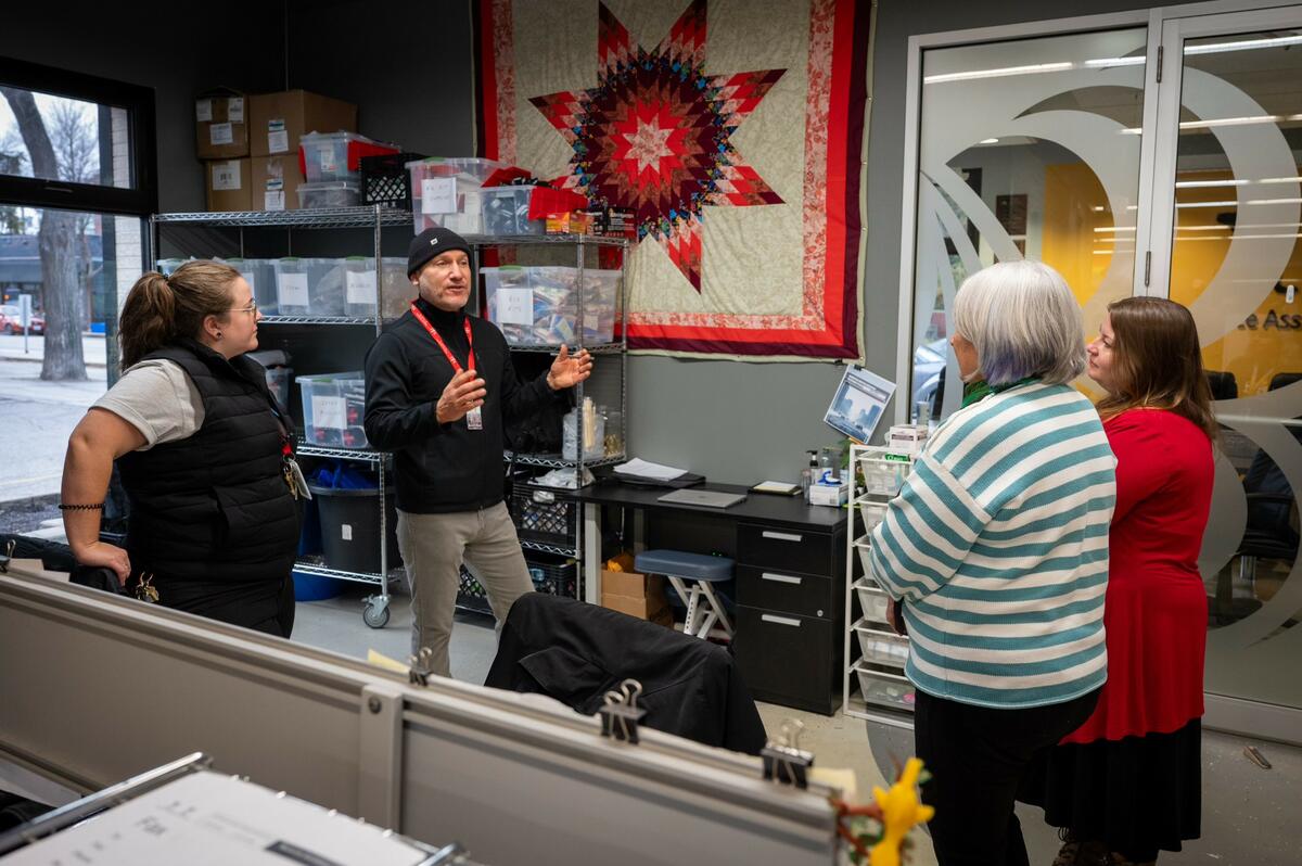 Governor General Mary Simon is seen from behind, standing and wearing a striped white and green sweater, while she is listening to a gentleman wearing a black beanie hat. Two other women are also listening.